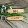 Two_wooden_bottle_openers_with_engraved_text_on_a_wooden_surface_with_greenery.