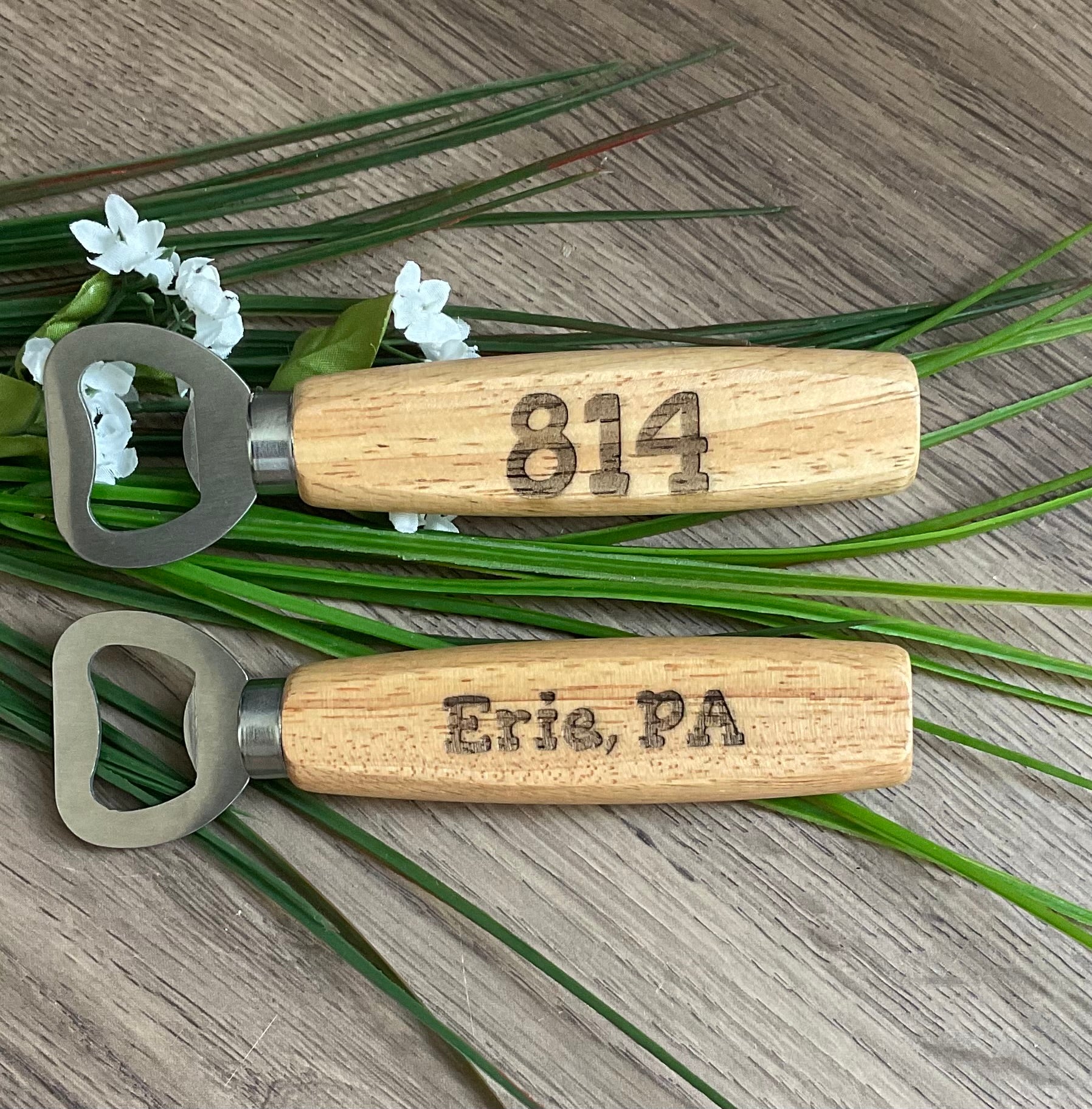 Two_wooden_bottle_openers_with_engraved_text_on_a_wooden_surface_with_greenery.